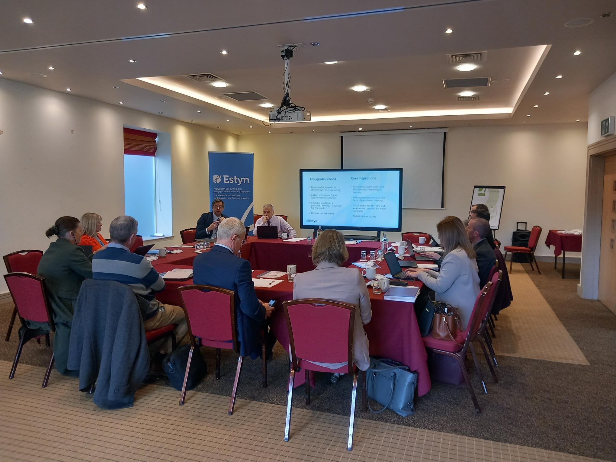 Attendees seated around a conference table in a meeting room at an Estyn event, with presentations displayed on a screen.