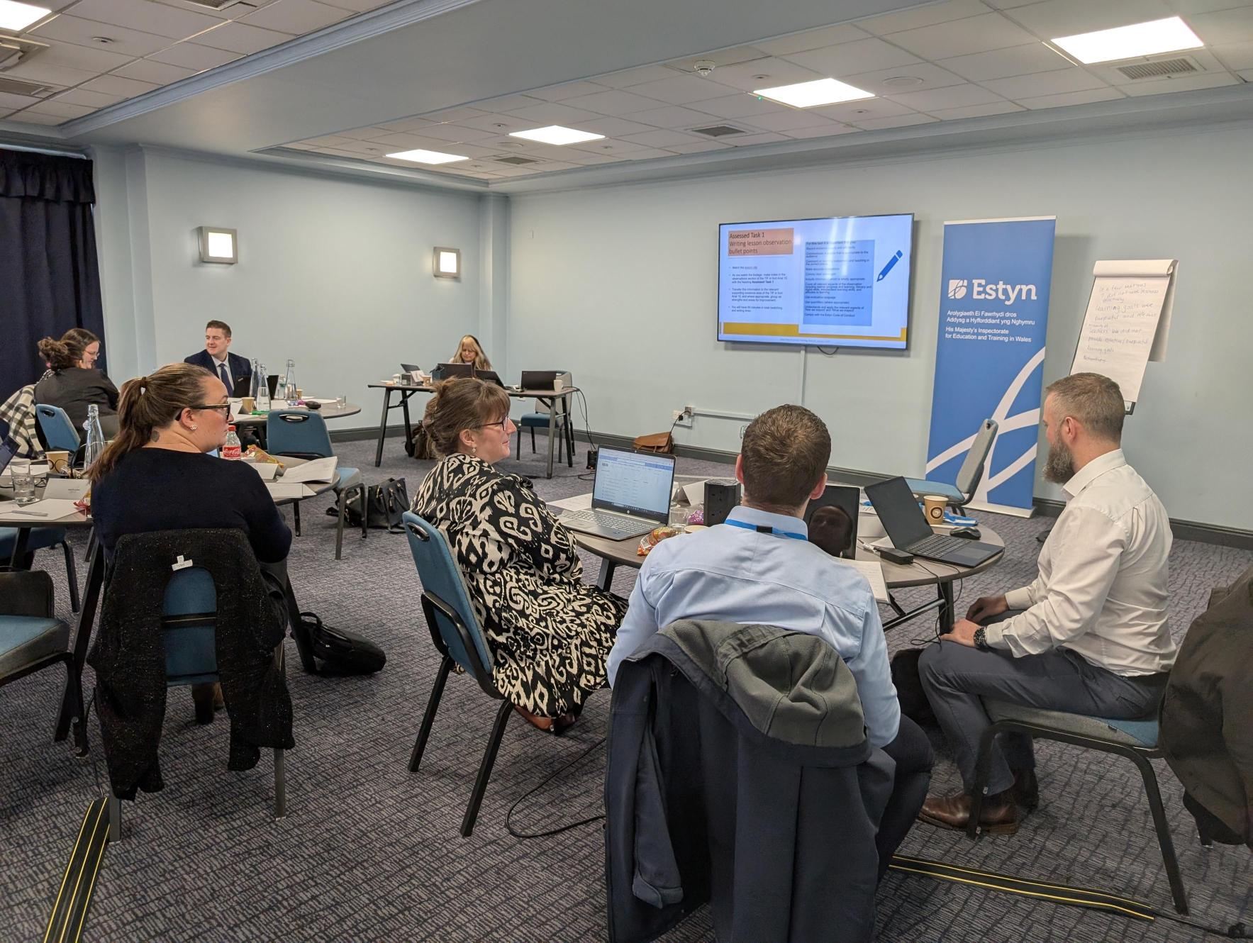 Group of professionals attending a workshop in a conference room with a presentation on screen and Estyn banners visible.