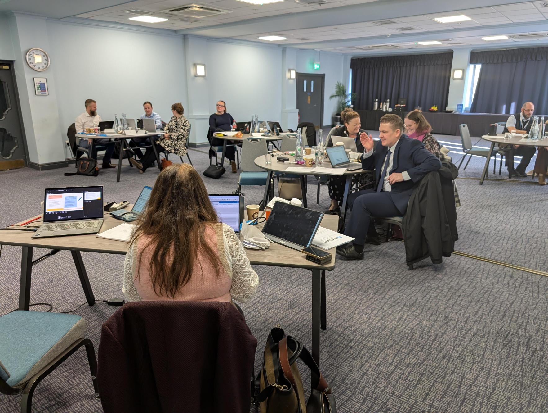Individuals seated at tables during a conference session, working on laptops and engaging in discussions.