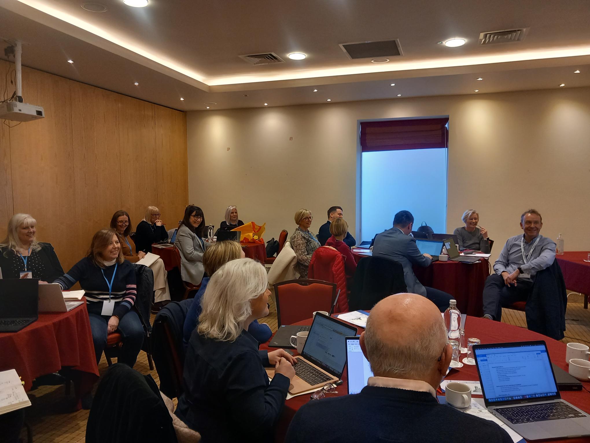 Participants seated at a conference room during a meeting, with some using laptops and others focusing on a presentation screen at the front.