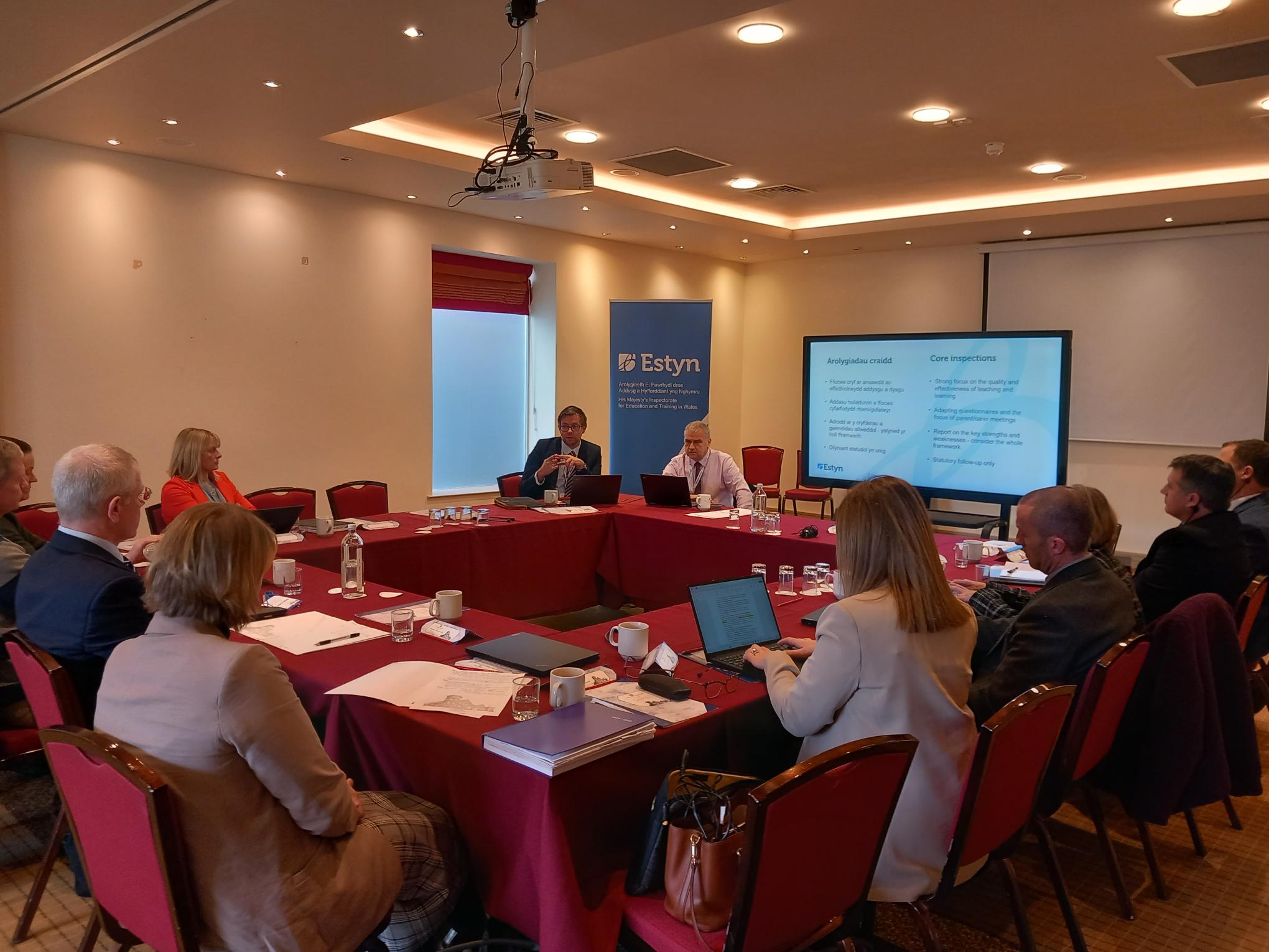 A business meeting in progress with participants seated around a table, facing a presentation labeled "Estyn." Two speakers are presenting in front of a screen displaying graphs and bullet points.