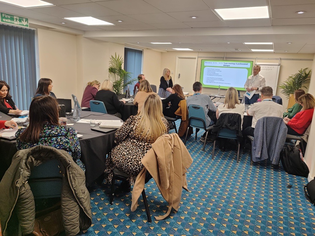A business workshop in a meeting room with attendees seated at tables listening to a presenter near a digital screen displaying a slideshow.