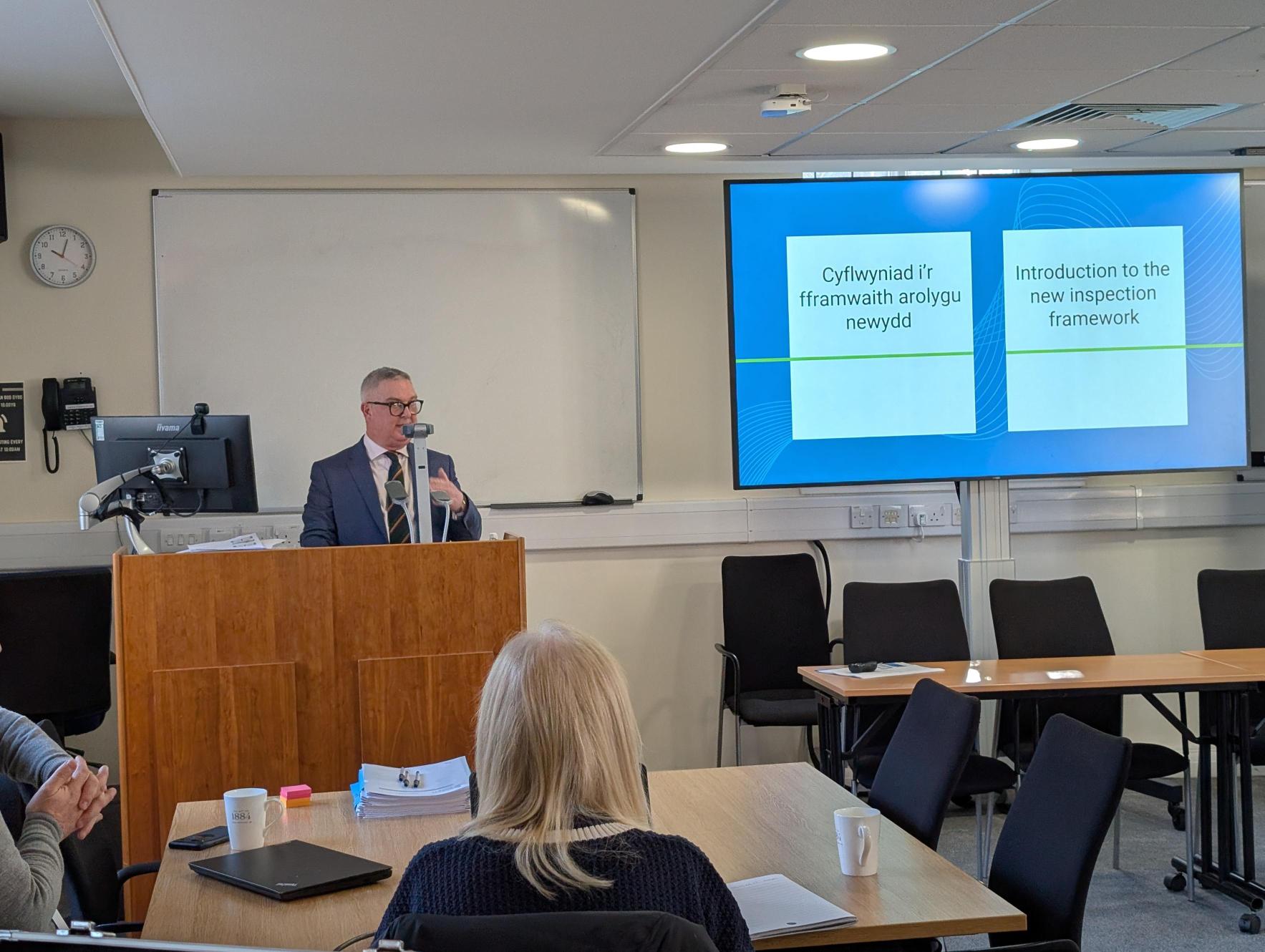 Person giving a presentation at a podium about the new inspection framework to an audience in a classroom. The presentation slide is bilingual, written in both Welsh and English.