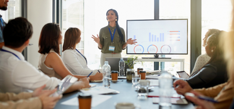 A professional presentation in progress, featuring a speaker at the front addressing a group of attendees seated around a conference table. The focus is on a screen displaying charts with percentages. The setting suggests a bright, modern office environment.