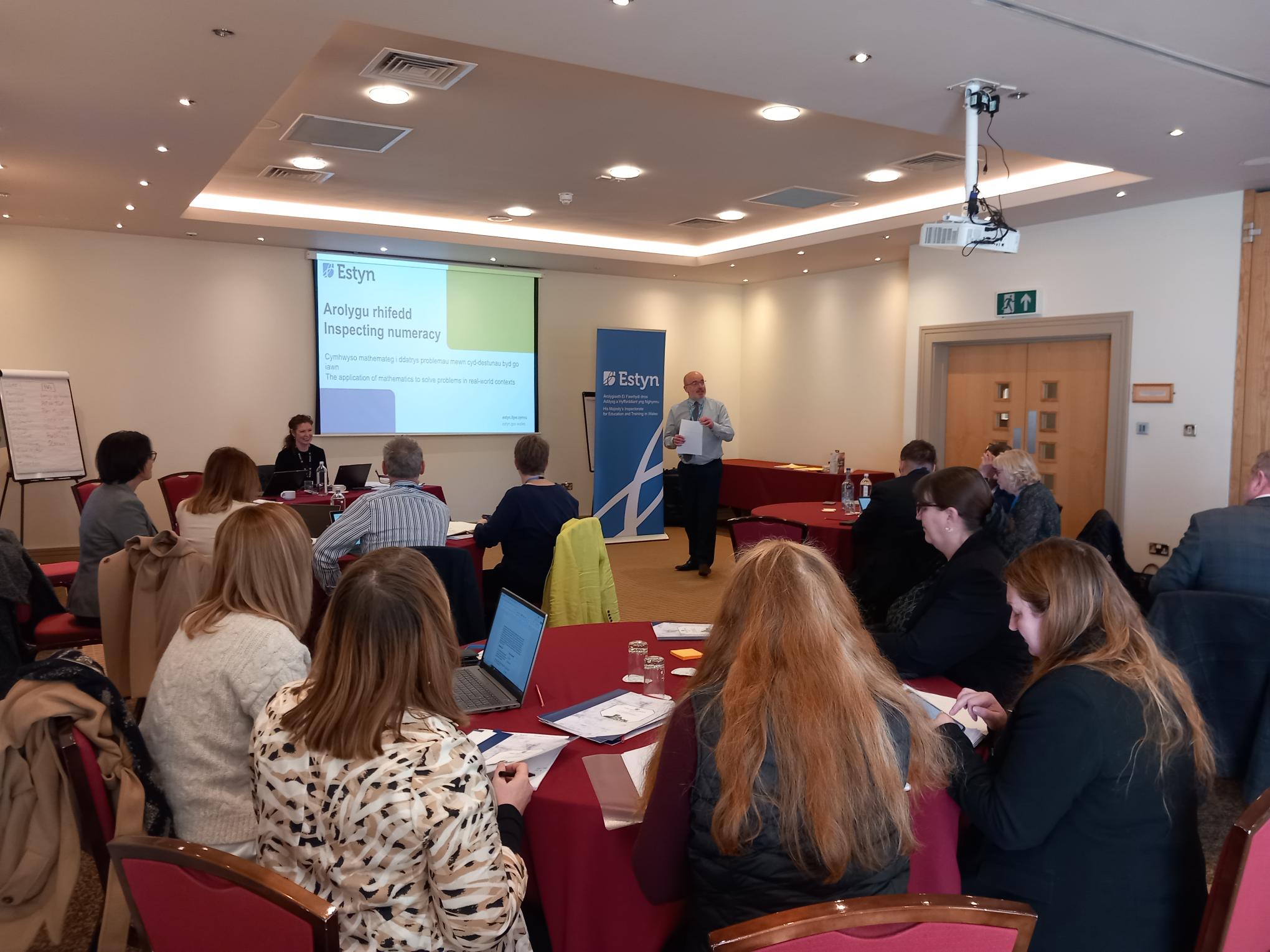 A professional meeting with participants seated at tables watching a presenter near a screen displaying a PowerPoint about "Inspecting numeracy" for Estyn.