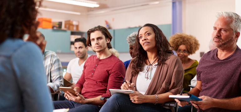 A group of adult students with notebooks sitting in a classroom, attentively listening to a speaker who is off camera.