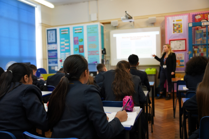 A classroom with students seated at desks listening to a teacher presenting a slide