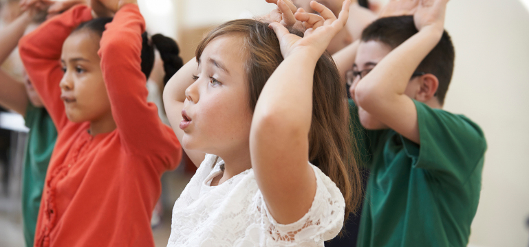 Children participating in a dance class, raising their arms overhead in a coordinated movement.