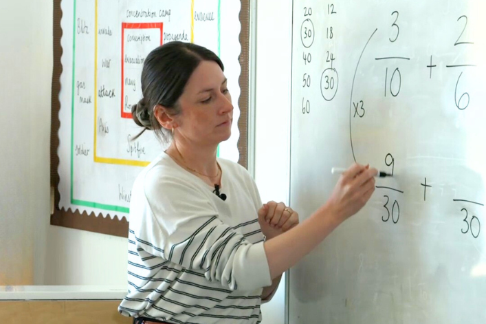 A teacher is writing a fraction addition problem on a whiteboard in a classroom.