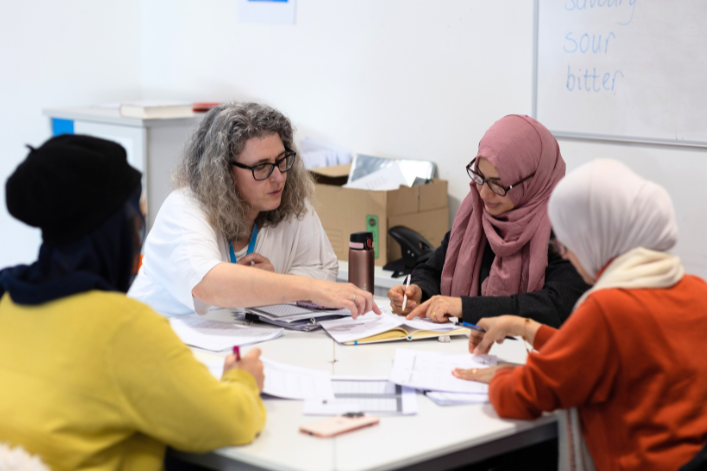 A group of adult students and a teacher discussing around a table in a classroom setting.