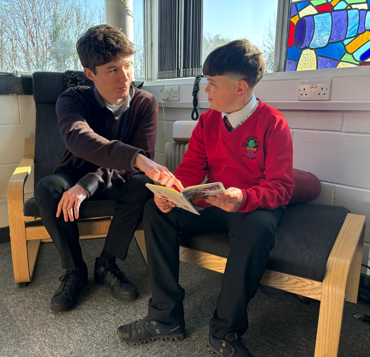 Two students, one in a school uniform, are engaged in reading a book together while sitting in a room with a stained glass window in the background.