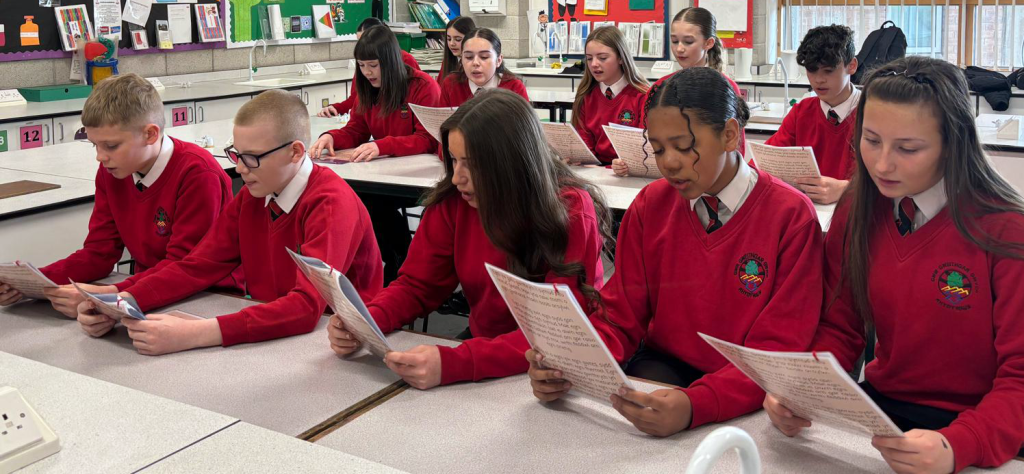 Students in school uniforms reading papers at desks in a classroom.