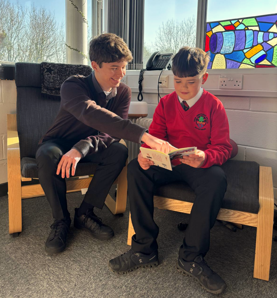 Two students, one in a brown sweater and the other in a red school uniform, are sitting by a window, reading a book together. A colorful stained glass window is visible in the background.
