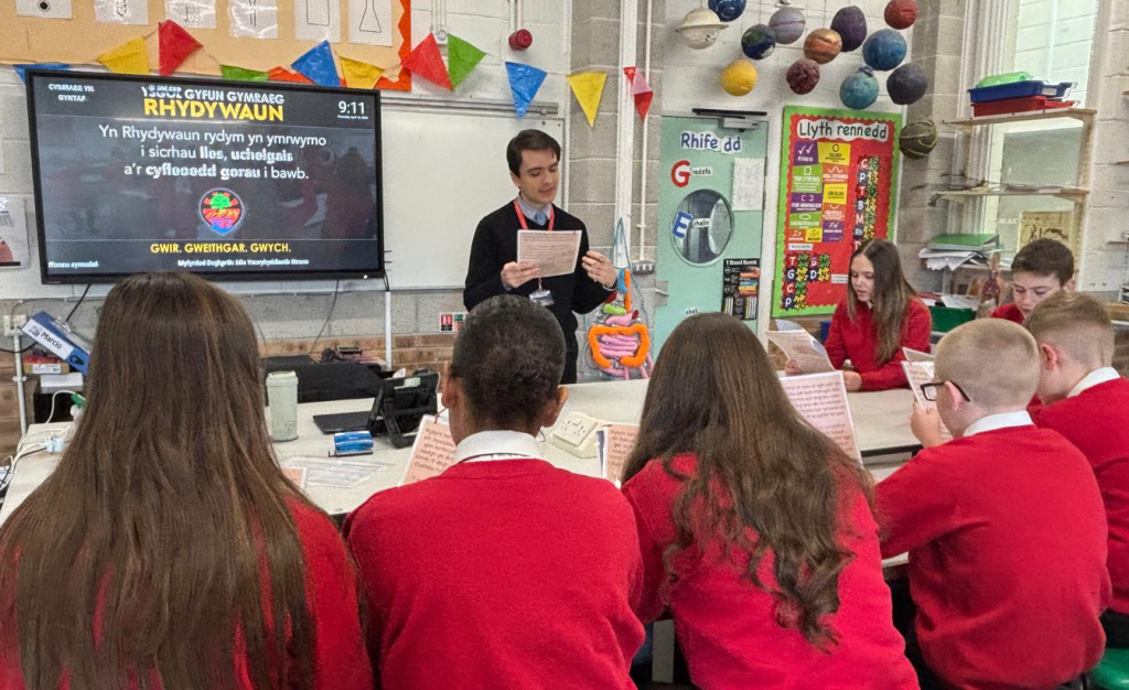 A teacher standing in front of a classroom, reading from a paper to students sitting at desks. The environment is colorful with educational decorations around.