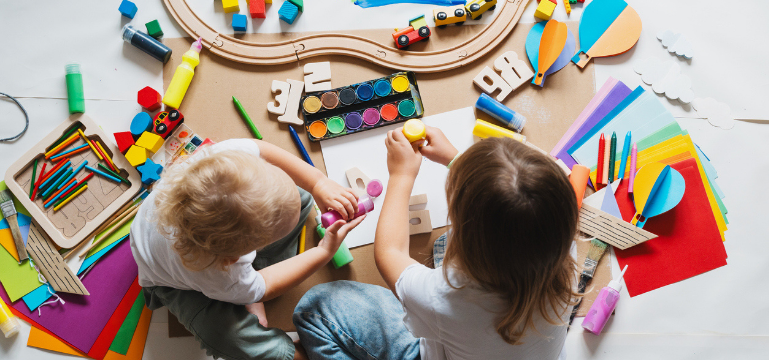 Two children are sitting on the floor, engaged in creative play with colorful craft materials and a wooden train track setup.