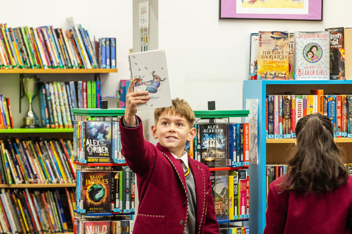 Child in a school uniform holding up a book in a library, with another child and bookshelves filled with books in the background.