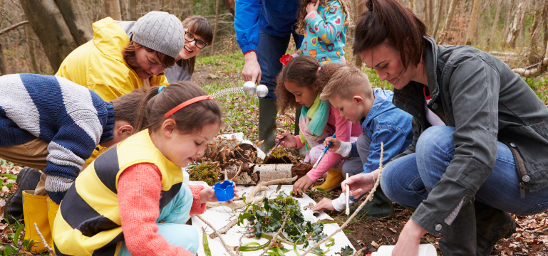 A group of adults and children engaging in a nature study outdoors, examining plants and insects together.