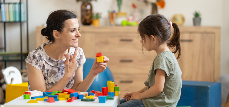 Two people, one adult and one child, are sitting at a table playing with colorful building blocks. They appear engaged and happy as they interact with each other. The background features a tidy room with a wooden shelf.