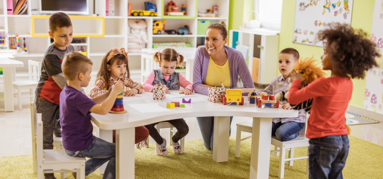 Children are seated around a curved table in a colorful classroom, engaging with educational toys. An adult is sitting with them, smiling. The room is bright, with shelves filled with various toys and books. One child stands near the table holding a stuffed toy.