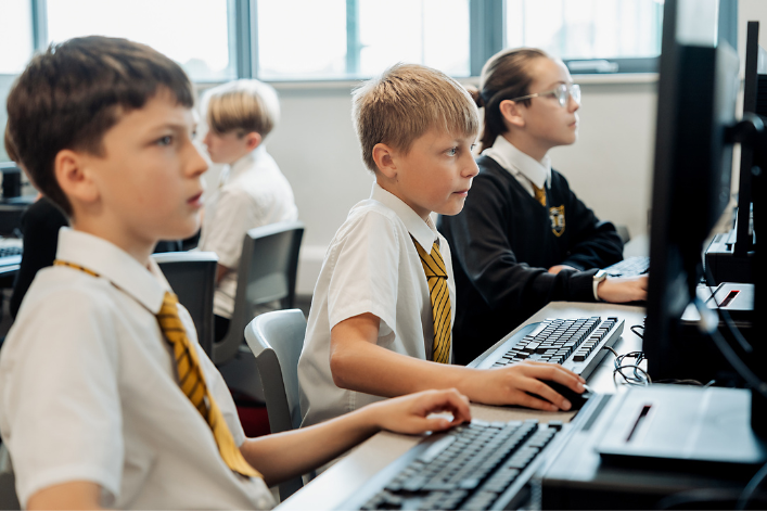 Several children in school uniforms sit at computer workstations in a classroom, each focused on a desktop computer. The room is bright with natural light from large windows in the background.