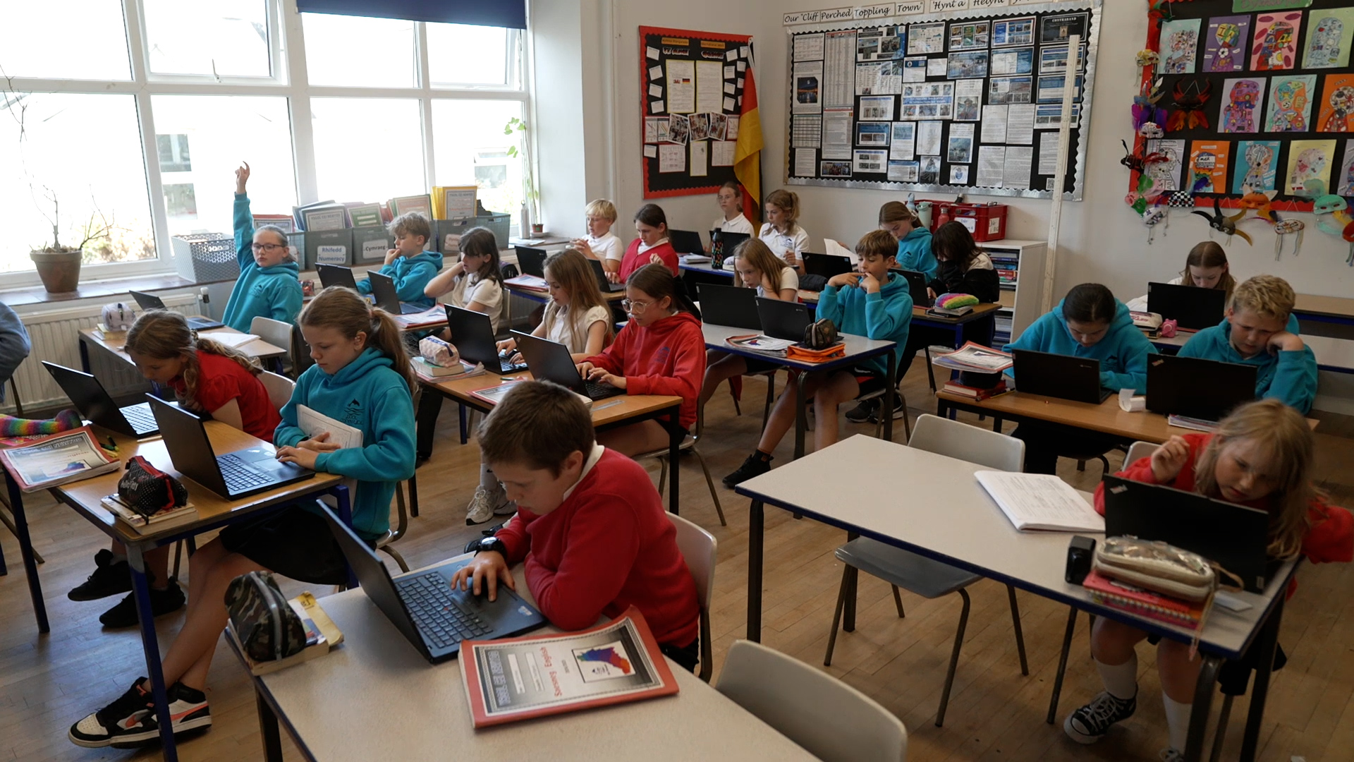 Dosbarth o fyfyrwyr ysgol gynradd yn eistedd wrth fyrddau unigol, pob un yn defnyddio gliniadur. Mae'r rhan fwyaf yn canolbwyntio ar eu sgriniau tra bod un myfyriwr yn codi llaw. Mae'r ystafell yn llachar gyda ffenestri mawr, ac mae'r waliau'n arddangos posteri addysgol a gwaith celf myfyrwyr.