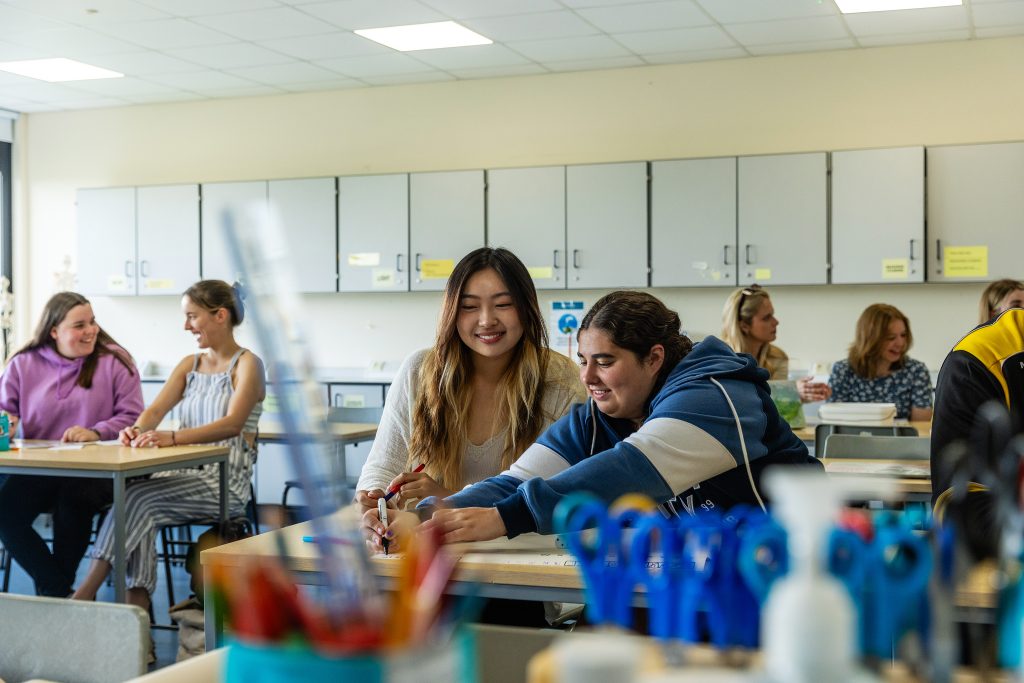 Students in a bright classroom working together at desks, with two in the foreground collaborating on a task while others chat and learn in the background.