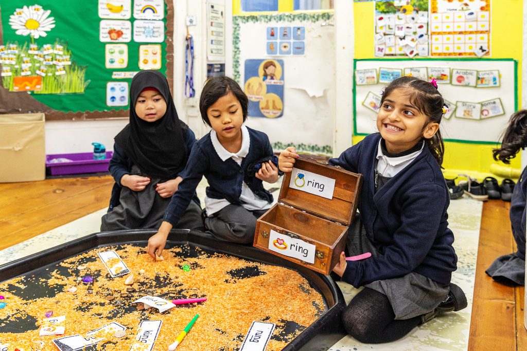 Young children in a classroom gathered around a tray of grains, exploring objects and word cards, while one child holds an open box labeled “ring” during a hands-on learning activity.