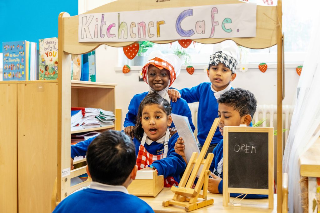 Plant yn chwarae rôl mewn caffi ystafell ddosbarth, yn gwisgo ffedogau a sgarffiau pen wrth weini cyd-ddisgyblion wrth til ffug o dan arwydd yn darllen “Kitchener Cafe”.