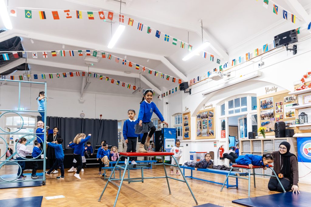 Plant yn cymryd rhan mewn gweithgareddau corfforol dan do mewn neuadd ysgol, yn dringo, yn cydbwyso, ac yn chwarae ar offer campfa tra bod staff yn goruchwylio yn y cefndir.