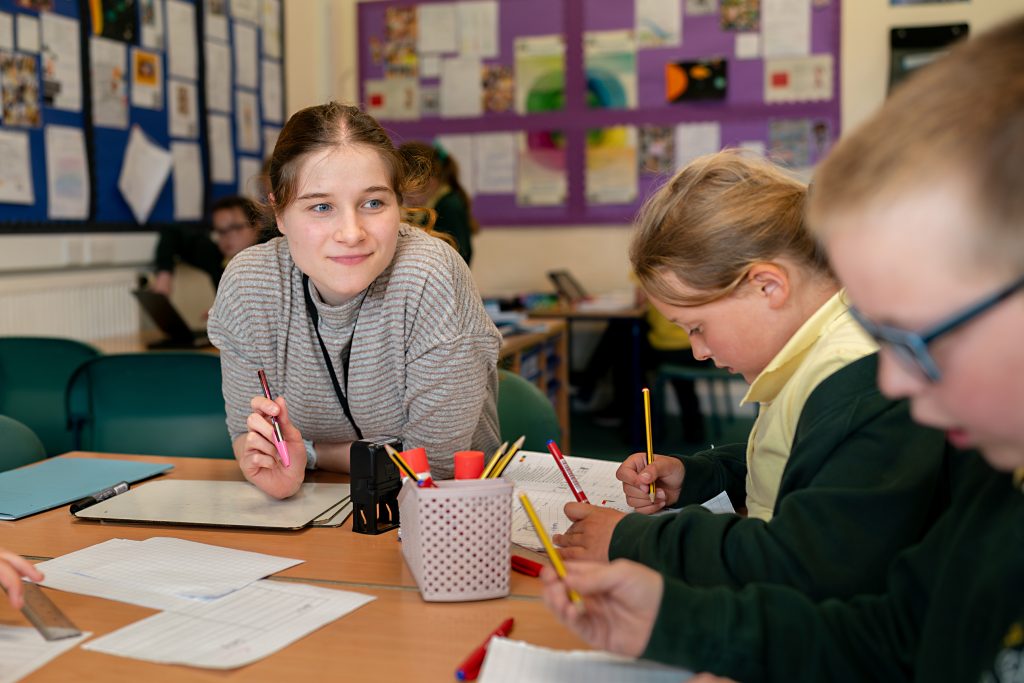 Athro yn cefnogi disgyblion ysgol gynradd wrth iddynt ysgrifennu a chwblhau gweithgareddau ystafell ddosbarth gyda'i gilydd wrth fwrdd wedi'i orchuddio â phensiliau a thaflenni gwaith.