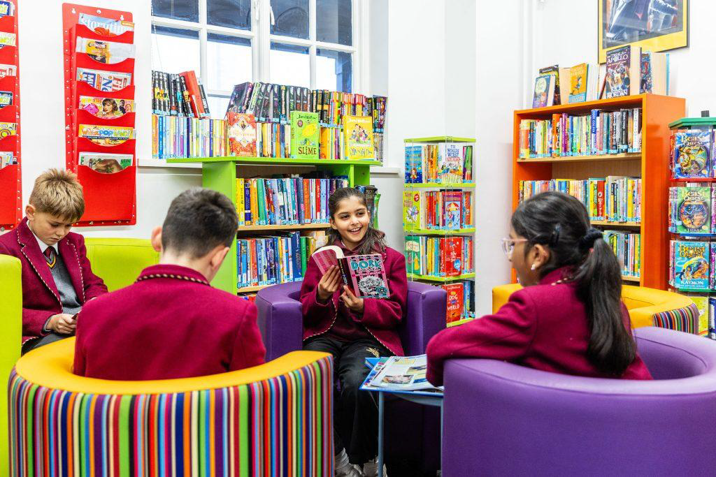Four individuals in red uniforms are seated in a library with colorful, round chairs. Bookshelves full of children's books, including titles like "Star Wars," are in the background. One person is holding an open book and smiling.