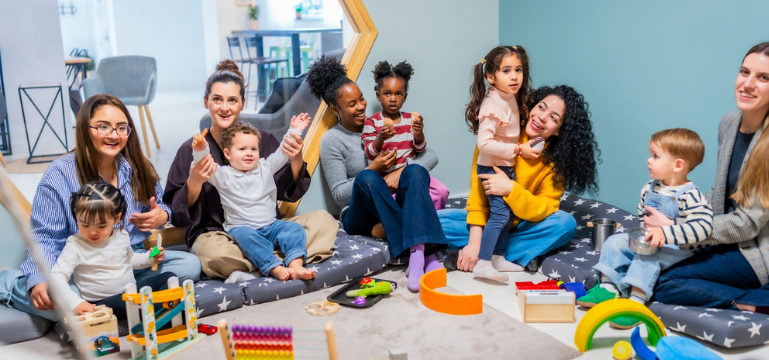 A group of young children and adults sitting on cushions in a playroom, holding toys and facing the camera, with colourful play equipment scattered on the floor.