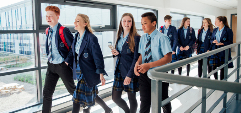 A group of secondary school students in uniform walking along a bright indoor corridor with large windows, some carrying bags and one holding a phone.