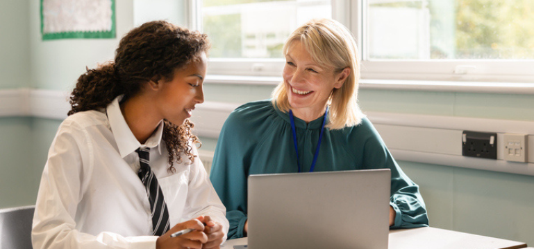 A student and an adult sitting at a table with a laptop, talking together in a classroom setting.