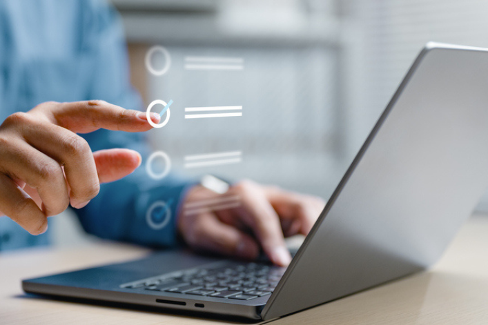 A close-up of a person using a laptop while pointing at a digital interface with selectable options displayed over the screen.