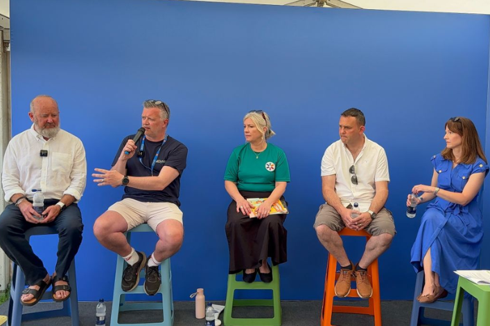Five people sit on colourful high stools on a stage in front of a blue backdrop during a panel discussion. One man speaks into a microphone while the others listen, each holding a water bottle or notes.
