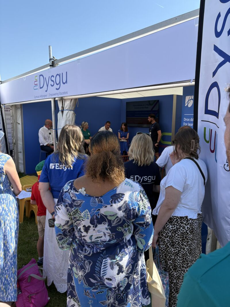 People stand outside the Dysgu education tent at an outdoor event, watching a panel discussion taking place inside. The blue-and-white branded tent features seated speakers on a small stage while attendees gather in the sunshine.