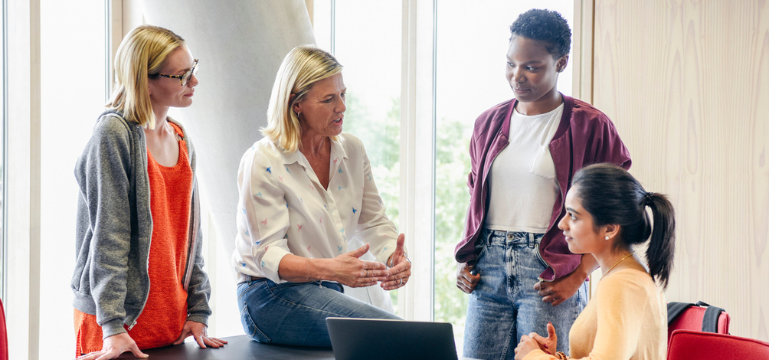 Four adults gathered around a laptop, discussing work together in a bright indoor setting.
