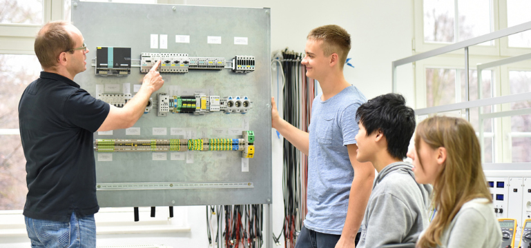 An adult pointing at an electrical control panel while three learners observe in a classroom or workshop setting.