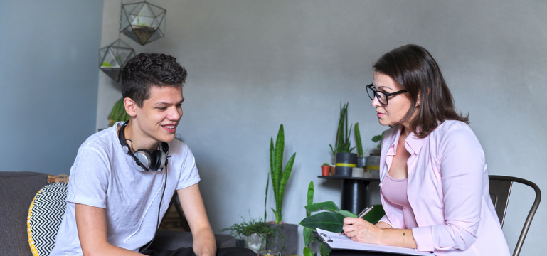 An adult and a younger person seated facing each other in a home setting, talking while one person writes notes on a clipboard.