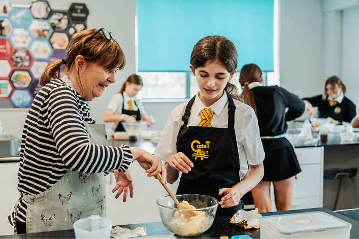 An adult and a student in a classroom kitchen preparing a mixture in a bowl, with other students working in the background.