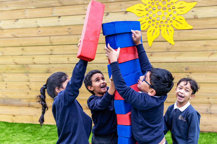 A group of children outdoors stacking large red and blue foam blocks together, with a wooden fence and a yellow decorative shape in the background.