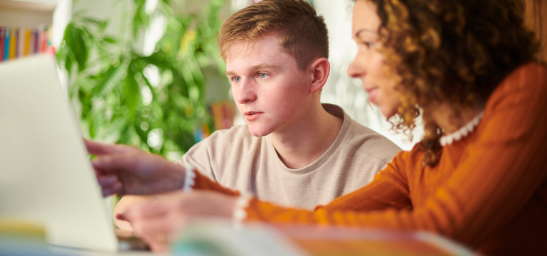 A student and an adult sitting at a table with a laptop, talking together in a classroom setting.