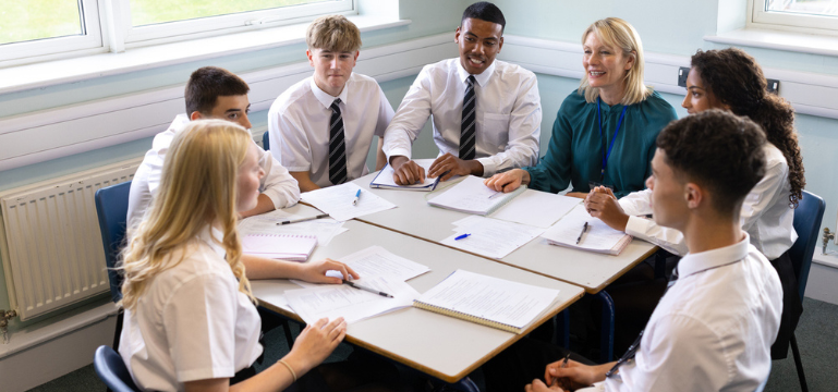 Students and teacher in a classroom discussion around a table.