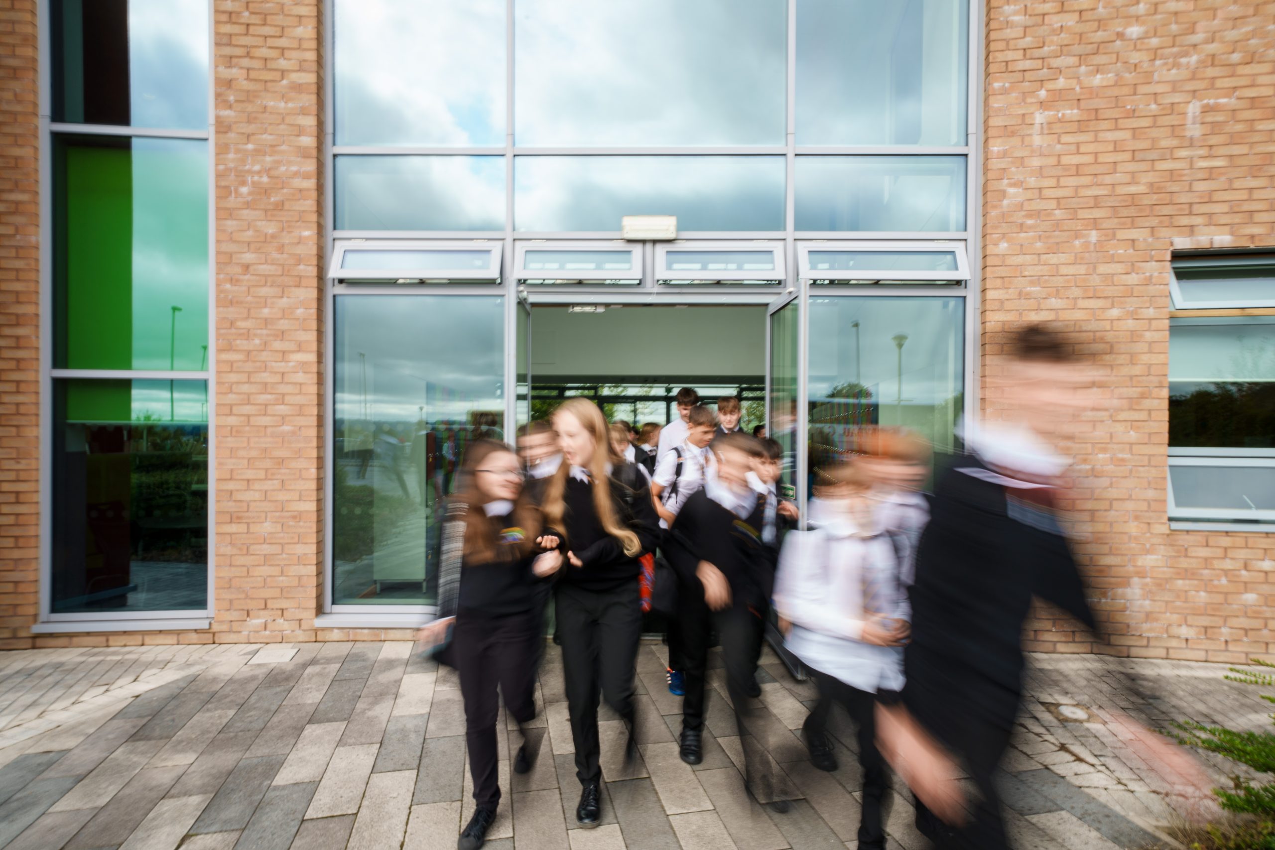 Pupils in school uniform walking out of a school building through open glass doors.