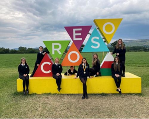 School children posing in front of a colourful work of art consisting of the word "CROESO" with each letter printed on a vibrant triangular block